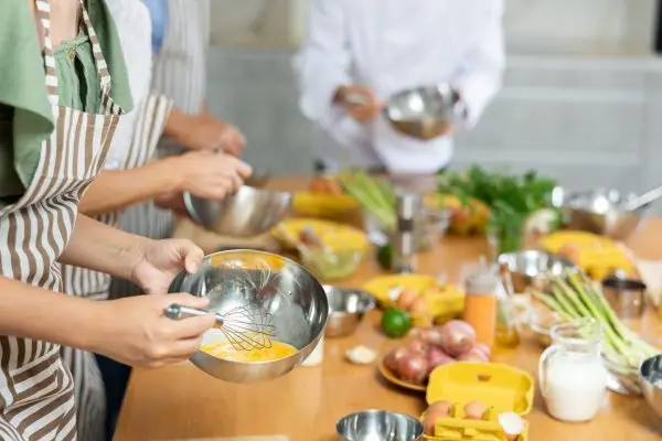 People participating group culinary class, engrossed in cooking process, beating up eggs with whisk following chef instructions, cropped shot..